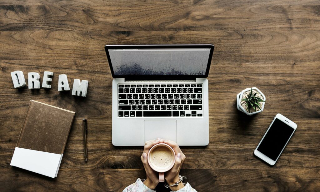 Na biurku jest laptop, na którym jest robiona strona na Wordpress, telefon, długopis, notesik i ręce Pani która trzyma kawę